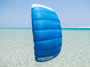 Blue kite labeled 'IGNITION' over clear water with a blue sky. IGNITION V3 - Trainer Kite ozone kids first kite portugal